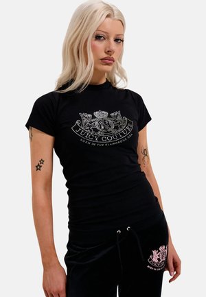 Jonge vrouw met blond haar draagt een zwart Juicy Couture T-shirt en bijpassende broek, en toont ster- en spinnen-tattoos op haar armen.