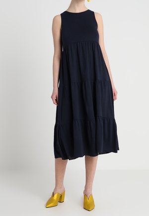 Vestido midi sin mangas azul marino con falda de volantes, combinado con mules amarillos de tacón ancho y punta en un persona de pie sobre un fondo blanco.