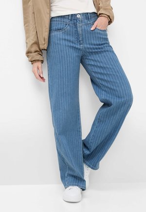 Kvinde iført lys brun jakke og hvid skjorte med blå, brede bukser med striber og hvide sneakers, poserer med den ene hånd i lommen.