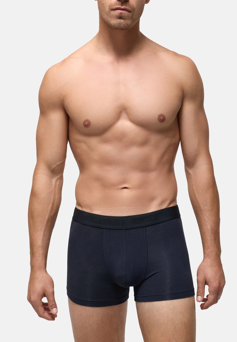 Muskelöser Mann trägt dunkelblaue Boxershorts vor einfachem hellgrauem Hintergrund, Torso und obere Oberschenkel sichtbar.