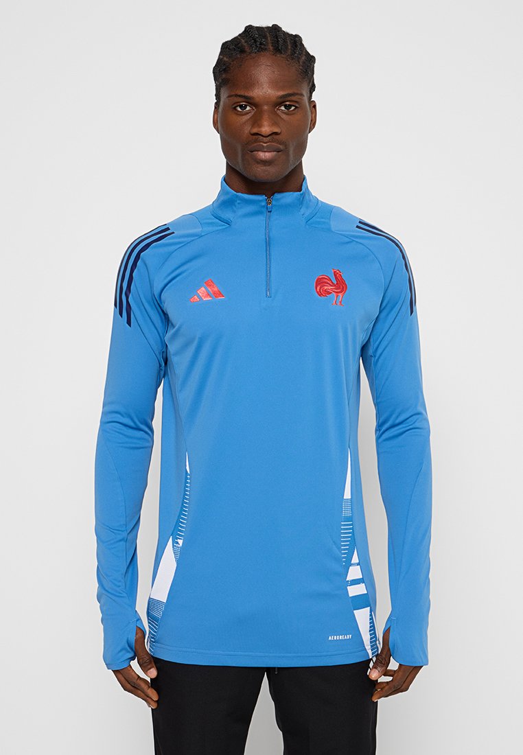 Jersey atlético azul con mangas largas, media cremallera, emblema de gallo rojo y franjas negras en los hombros, con acentos geométricos blancos.