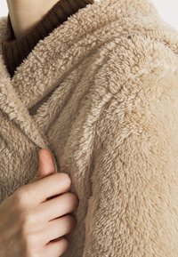 Beżowy kardigan z kapturem, wykonany z miękkiego, teksturowanego polaru z widocznym szwem i ręką chwytającą materiał, podkreślającą jego przytulność.