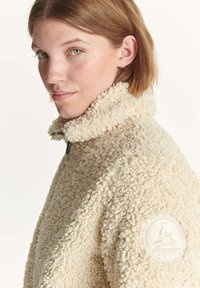 Giacca in pile beige con colletto alto, superficie testurizzata e una toppa circolare con logo sulla manica sinistra. Presenta una chiusura con zip.