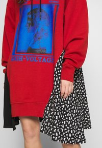 Personne portant une robe sweat-shirt rouge avec un motif bleu et le texte "HIGH-VOLTAGE", associée à une jupe fleurie noire et blanche.