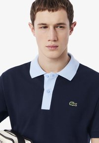 Marinblå polo med ljusblå krage, två knappar och broderad grön krokodil-logo på vänster bröst. Blandad bomullstextil med texturerad yta.