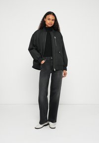 Μαύρο μπουφάν τύπου bomber με μπροστινό φερμουάρ, ριμπ μανσέτες και πλαϊνές τσέπες, σε συνδυασμό με ψηλόμεσα σκούρα γκρι τζιν και λευκά παπούτσια.