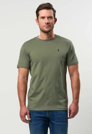 Man met kort bruin haar, die een olijfgroen T-shirt met korte mouwen en een blauwe spijkerbroek draagt, staand tegen een effen lichte achtergrond.