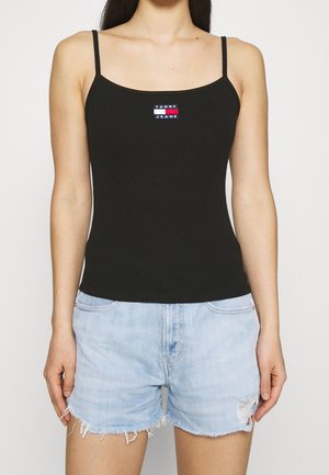 Femme portant un débardeur noir à fines bretelles avec le logo Tommy Jeans et un short en jean bleu clair usé sur un fond uni.