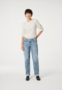 Lichte crèmekleurige, geribde trui met pofmouwen; bedrukte lichtblauwe jeans met zonnebloemen; zwarte loafers; neutrale achtergrond.