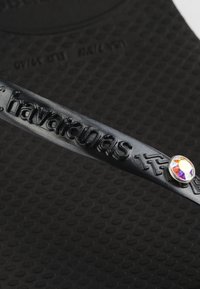 Μαύρο λουράκι από καουτσούκ για σαγιονάρα με ανάγλυφη την επιγραφή «havaianas» και μικρό στρογγυλό κόσμημα προσαρτημένο κοντά στην πλευρά.