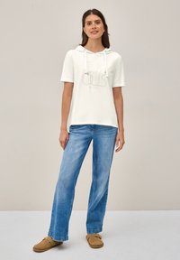 Haut à capuche léger blanc avec un design de logo, manches courtes et cordons de serrage. Associé à un jean bleu à jambes larges et des chaussures à enfiler marron.