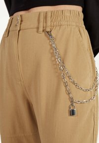 Beige cargobroeken met een elastische tailleband hebben een zilveren ketting met een hangslot bevestigd, wat een gedetailleerd accessoire element creëert.