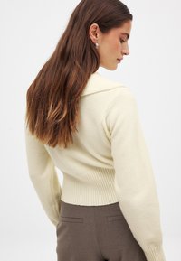 Hellgelbes, geripptes Strickoberteil mit weitem Kragen und langen Ärmeln, kombiniert mit braunen, figurbetonten Hosen, die einen strukturierten Stoff zeigen.