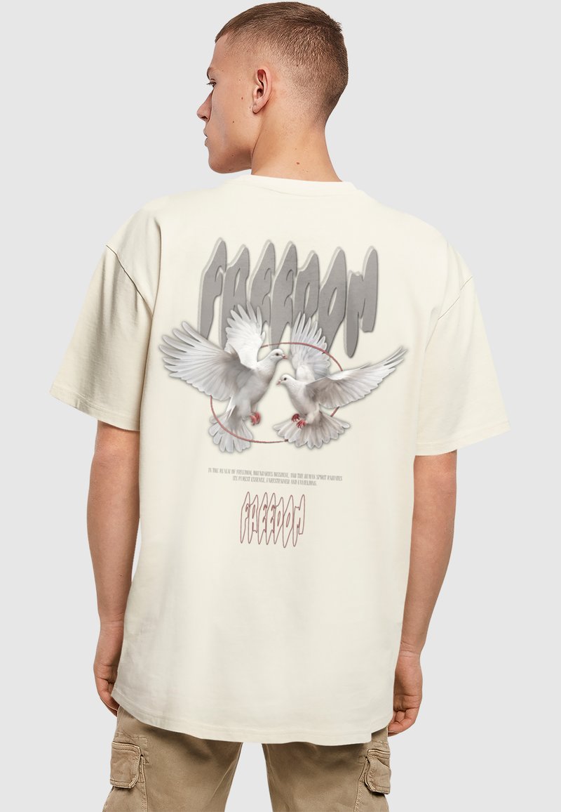 Beige over-size t-shirt med en grafisk design af to duer og ordet "FREEDOM" i stiliseret tekst på ryggen.