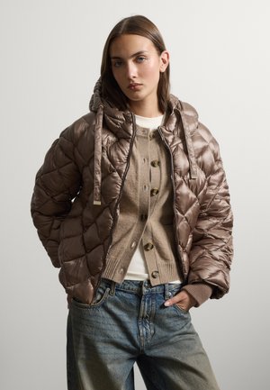 Young woman wearing a shiny brown quilted jacket with hood, beige buttoned cardigan, white top, and loose blue jeans, standing with one hand in pocket.