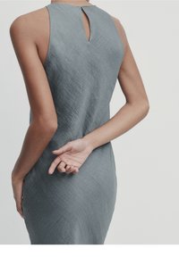 Robe sans manches grise en tissu texturé, avec une fente à l'arrière. Le mannequin a une main derrière le dos.