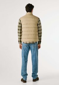 Gilet imbottito beige con cuciture orizzontali, sopra una camicia a quadri verde. Jeans in denim blu e stivali marroni completano l'outfit.
