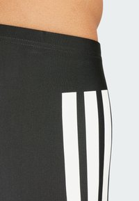 Pantaloni sportivi neri con una texture liscia, caratterizzati da tre strisce bianche verticali su un lato. Vita elasticizzata visibile.