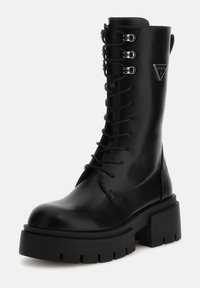 Botte noire en cuir montant jusqu'au genou avec une semelle plateforme noire épaisse, dotée de lacets à l'avant, d'accents en métal et d'un détail de logo triangulaire.