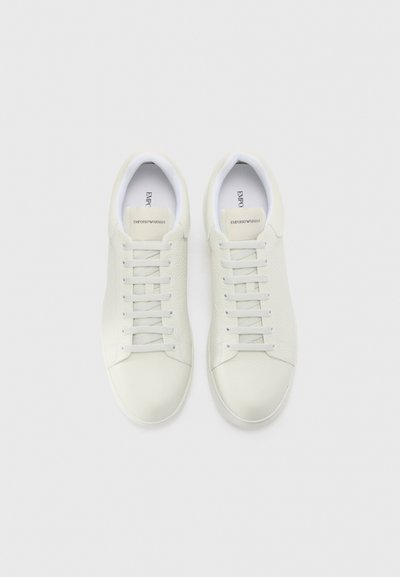 Baskets blanches en cuir avec une finition texturée, présentant une pointe arrondie, six œillets et un branding minimaliste sur la languette.