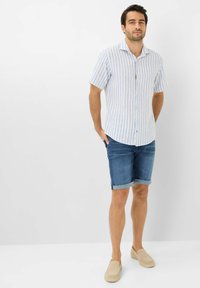 Camicia a maniche corte a righe in azzurro chiaro e bianco, abbinata a pantaloni corti in denim blu e scarpe slip-on beige. Chiusura semplice con bottoni davanti.