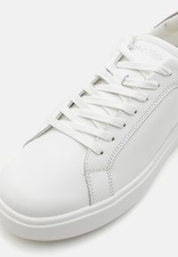 White leather sneaker with flat white laces, smooth texture, rounded toe, subtle stitching details, and a low-profile rubber sole.