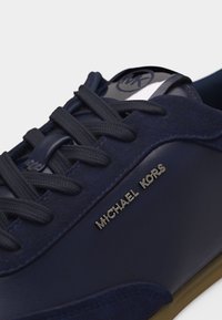 Σκούρο μπλε αθλητικό παπούτσι Michael Kors με κορδόνια στο ίδιο χρώμα, λεπτομέρειες από σουέτ και δέρμα, και μεταλλικό λογότυπο της μάρκας στο πλάι.