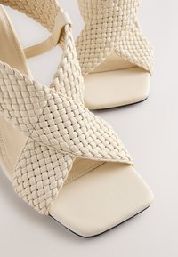 Sandales à talons tissées beiges avec un bout carré. Caractérisées par des brides tressées et une semelle en cuir lisse. Tige texturée avec des coutures détaillées.