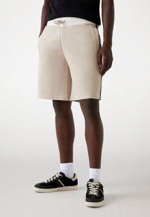 Beige athletic shorts with an elastic waistband and drawstring, featuring side pockets and contrasting black side stripes. Black sneakers with white accents.