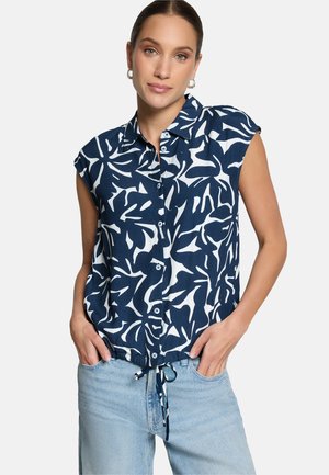 Femme portant une chemise sans manches bleu marine et blanche à motifs abstraits de feuilles, boutonnée et nouée à la taille, avec un jean bleu clair.