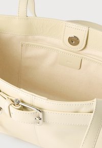 Δερμάτινη τσάντα χειρός σε εκρού χρώμα με λεπτομέρεια με αγκράφα, ανοιχτό πάνω μέρος που δείχνει το μπεζ ύφασμα εσωτερικά και εσωτερική θήκη για μικροαντικείμενα.