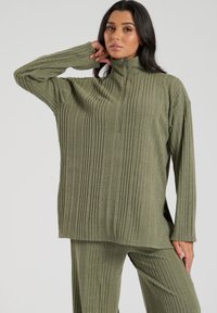 Olijfgroen fleece top met een hoge kraag, geribbelde textuur en een halve rits. Heeft een losse pasvorm met zijsplitten aan de onderkant.