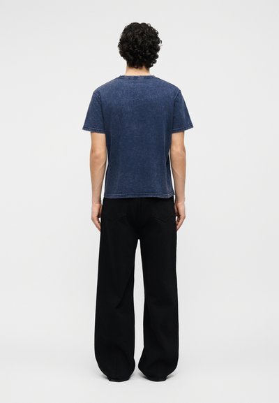 Άνδρας με σγουρά μαλλιά που φοράει ξεθωριασμένο μπλε κοντομάνικο μπλουζάκι και φαρδιά μαύρη παντελόνια, στέκεται με την πλάτη προς το μέρος μας σε απλό φόντο.