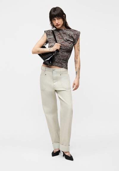 Νεαρή γυναίκα με τατουάζ, που φοράει αμάνικο μπλουζάκι με σχέδια, μπεζ παντελόνι με ψηλή μέση, μαύρα τακούνια και κρατά μια μικρή μαύρη τσάντα.