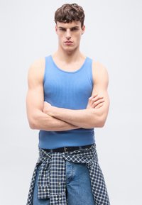 Νέος άνδρας που φορά μπλε αμάνικο πουκάμισο και τζιν, με ένα καρό πουκάμισο δεμένο γύρω από τη μέση του, στέκεται με σταυρωμένα τα χέρια.