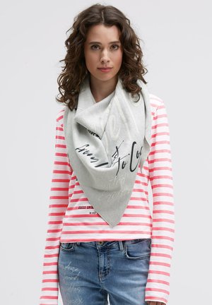 Frau mit lockigem Haar, die einen grauen Schal trägt, ein pink-weiß gestreiftes Langarmshirt und eine Jeans mit Schriftzug auf dem Oberschenkel.