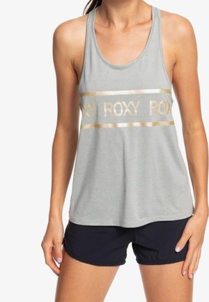 Kobieta w szarym bezrękawniku z napisem "ROXY" w kolorze złotym na piersi oraz w czarnych sportowych szortach, stojąca na białym tle.