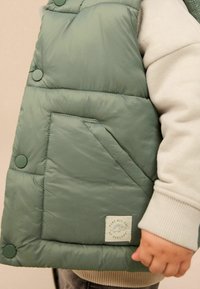 Gepolsterte Weste in gedämpftem Grün, mit Druckknöpfen und einer Fronttasche. Weicher Stoff mit dezentem Glanz, verziert mit einem kleinen Logopatch.