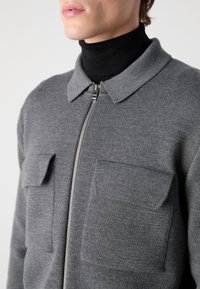 Šedá bunda na zip s límečkem, vyrobená z měkké pletené látky. Má dvě přední našité kapsy a přiléhavý střih.