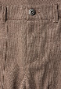 Tissu texturé brun de pantalons avec un motif en chevrons. Présente un bouton en métal noir et des coutures visibles le long de la taille.