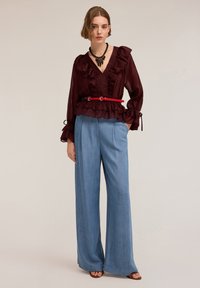 Blusa bordeaux a volant con maniche lunghe e scollatura a V, abbinata a pantaloni a gamba larga blu chiaro. Una cintura rossa segna la vita.