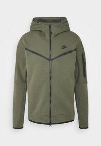 Olivgrön Nike-jacka med huva, svart dragkedja, svarta diagonala ränder och zip-ficka på vänster ärm.