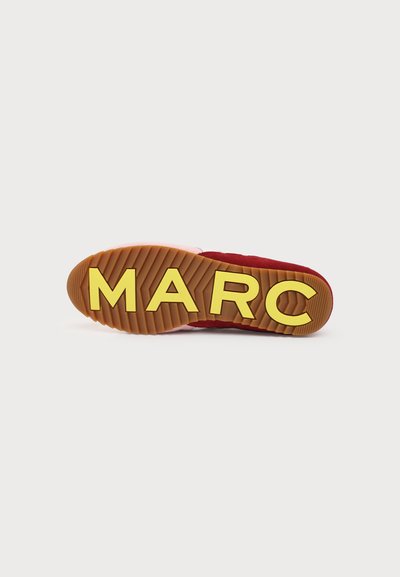 Semelle de chaussure marron avec de grandes lettres jaunes formant "MARC,", motif de semelle rainuré, dessus rouge et rose partiellement visible.