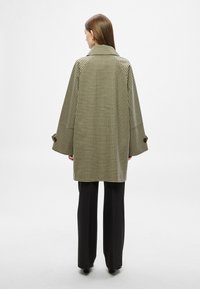 Manteau à carreaux en tissu noir et blanc, avec un large col et des manches surdimensionnées. Porté avec un pantalon noir ajusté.