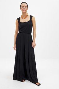 Moteris vilkinti juodą be rankovių viršų, plačias juodas klostuotas kelnes, gintarinę masyvų karolį ir juodus kvadratinius batelius ant baltos fono.