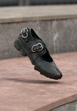 Czarny but z rozdzielonym palcem, z dwoma srebrnymi klamrami, grubą podeszwą i perforowanymi detalami, unoszący się nad kamienną powierzchnią.