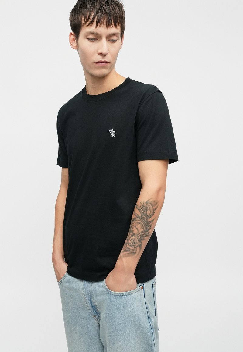 T-shirt noir en coton, avec un petit motif sur la poitrine gauche, associé à un jean en denim délavé. Encolure ronde simple, manches courtes.