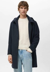 Abrigo de lana azul marino con frente de botones y cuello ancho, llevado sobre un suéter crema de punto trenzado y jeans de mezclilla claros.