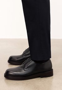 Pantofi negri din piele, cu vârfuri rotunjite, talpă groasă și șireturi, asortați cu pantaloni închisi la culoare, pe o suprafață netedă de culoare deschisă.