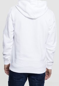 Persona che indossa un semplice hoodie bianco e jeans blu scuro, vista da dietro su uno sfondo neutro.